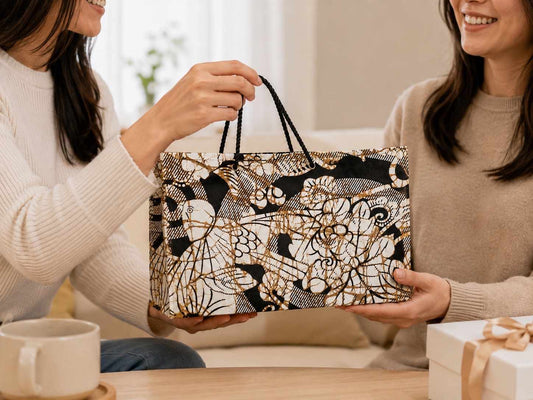 Two women holding a cream, black, and gold decorative bag with intricate patterns in a cozy indoor setting.
