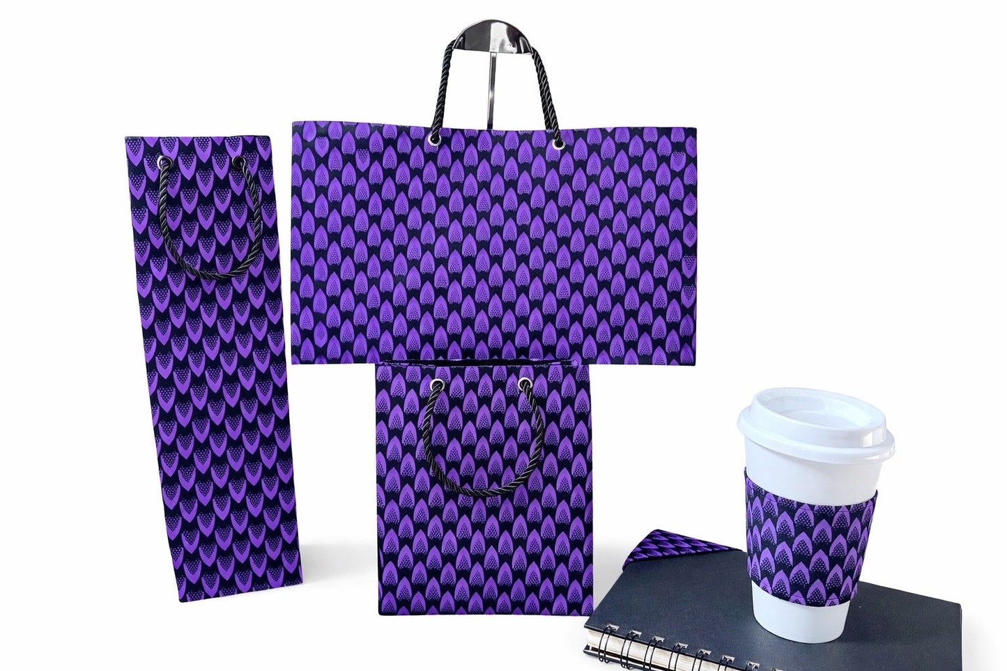 Purple and black patterned gift bags, spiral notebook with corner bookmark and a coffee cup with a cup sleeve on a white background