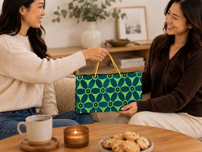 Two women sitting at a table with a green, dark green, and gold patterned gift bag, coffee, and cookies.