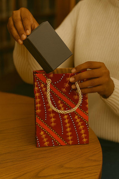 Person holding a small box over a decorative maroon, orange, black, & white  bag with intricate patterns on a wooden surface.