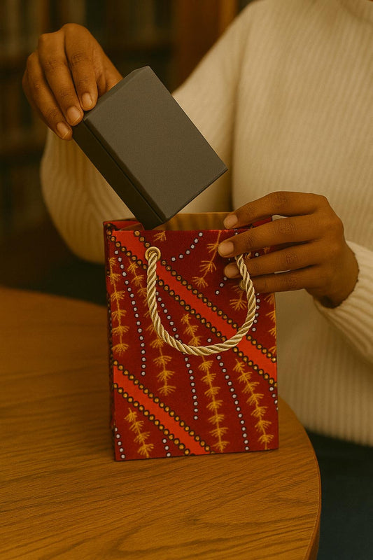 Person holding a small box over a decorative maroon, orange, black, & white  bag with intricate patterns on a wooden surface.