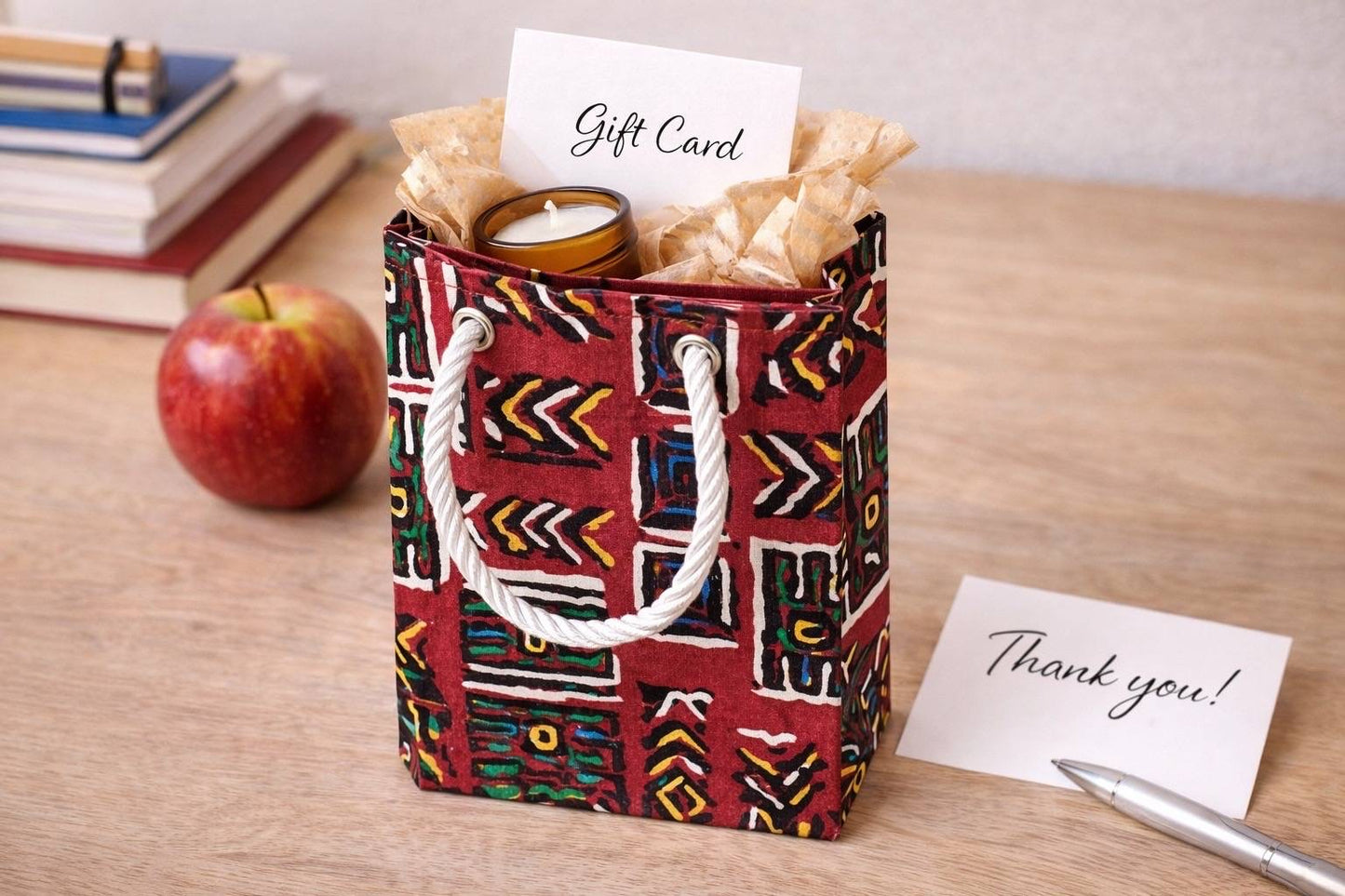 Decorative maroon, orange, blue, green, black, & white gift bag with a candle, card, and apple on a wooden surface
