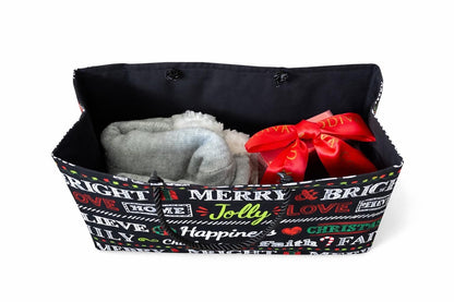 Black, red, green, and white gift bag with festive text and gray socks and coffee mug with chocolates and a red bow inside on a white background