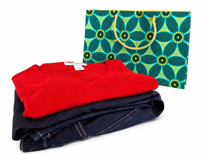 Folded red shirt and blue jeans with a patterned green, dark green, and gold gift bag on a white background