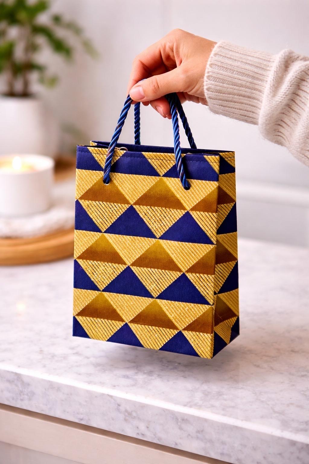 Hand holding a geometric patterned gold, dark blue, & brown gift bag with blue and gold design on a marble surface.