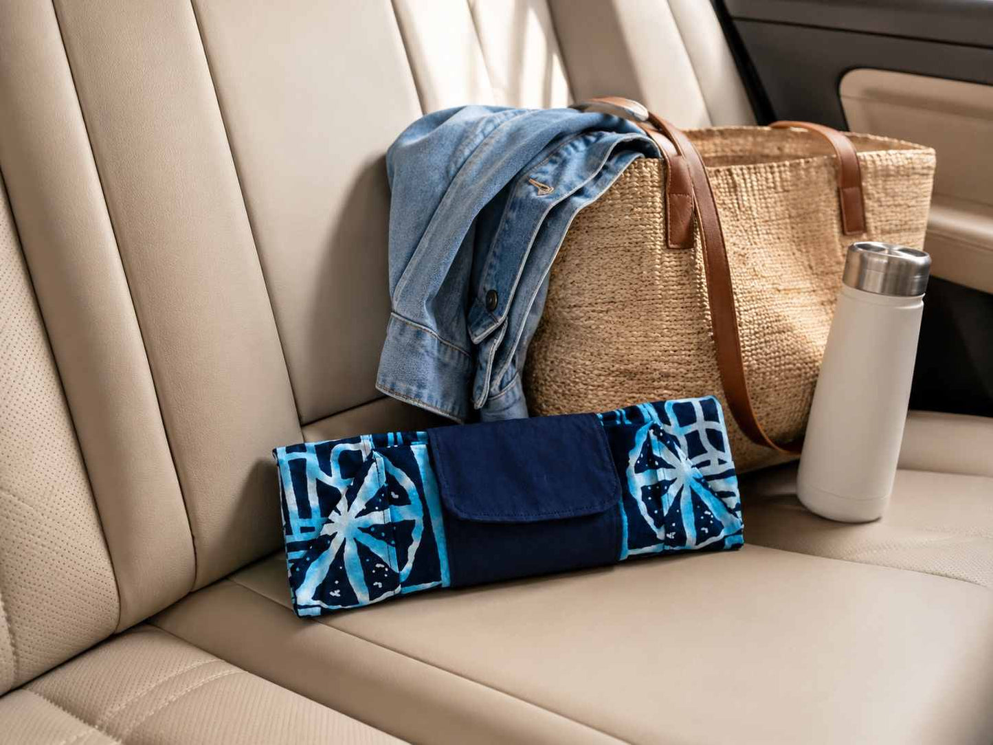 Car interior with a blue and white patterned folded in clutch position market bag, denim jacket, woven bag, and white tumbler.