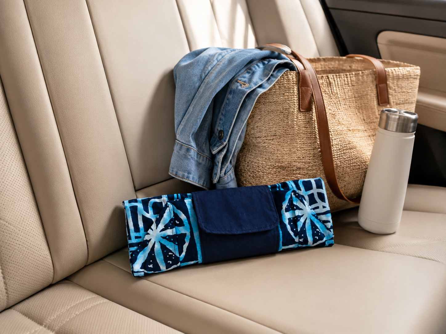 Car interior with a blue and white patterned folded in clutch position market bag, denim jacket, woven bag, and white tumbler.