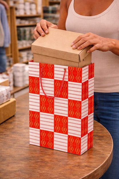 Person holding a shoe box over a decorative red, white, and gold gift bag with a checkered pattern on a wooden surface.