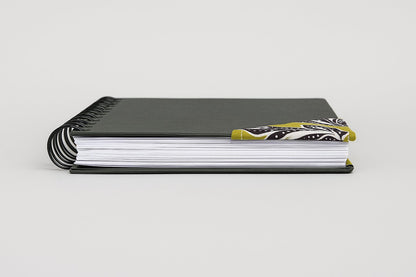Black notebook with a corner bookmark in green, brown, & white accents on a gray background