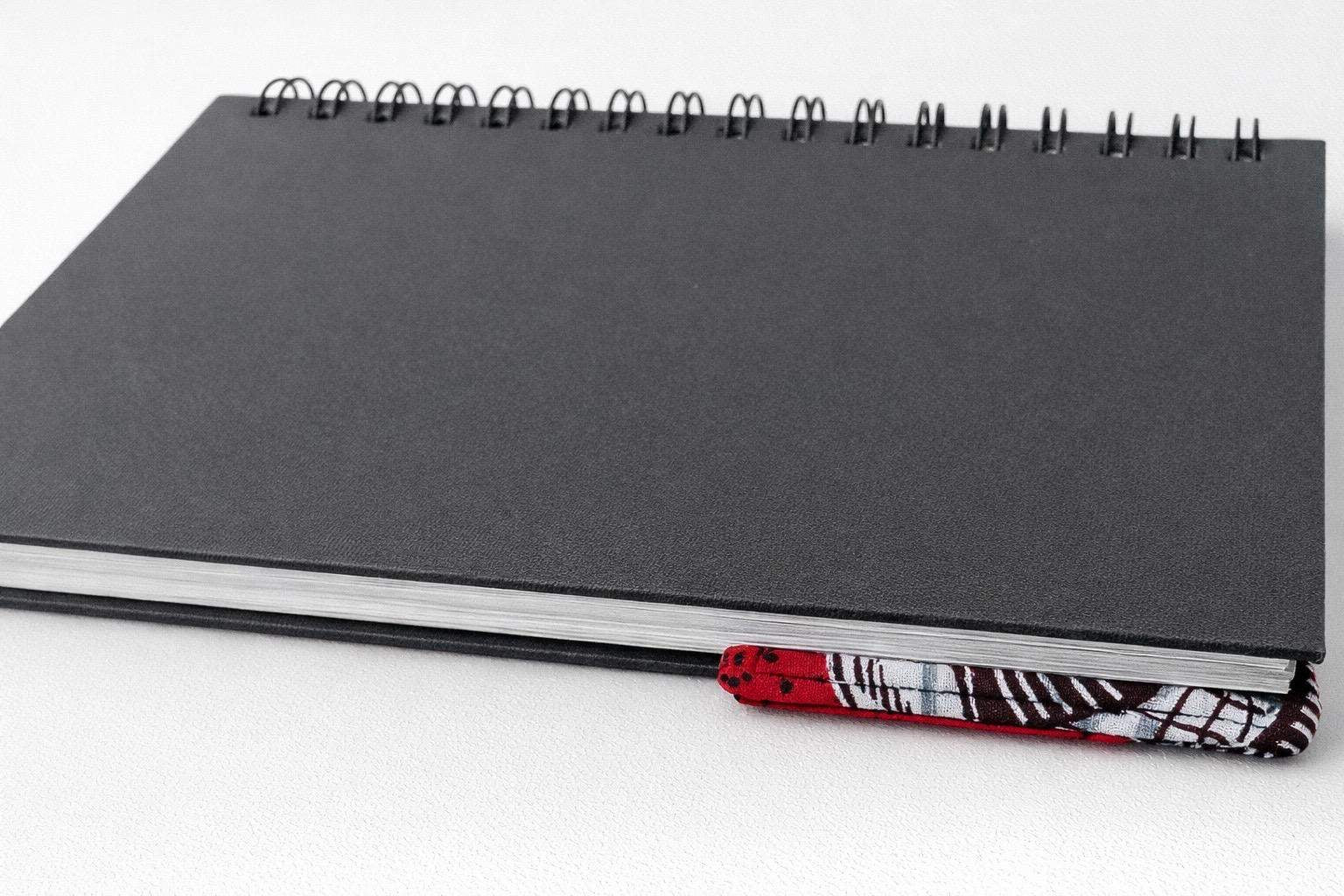 Black notebook with a corner bookmark in red, brown, & white accents on a gray background