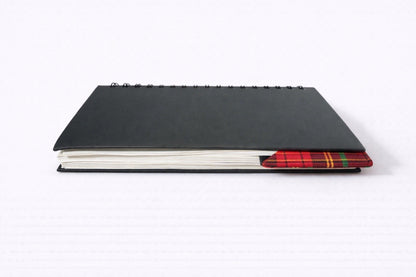 Black notebook with a black corner bookmark in red, green, & gold accents on a gray background