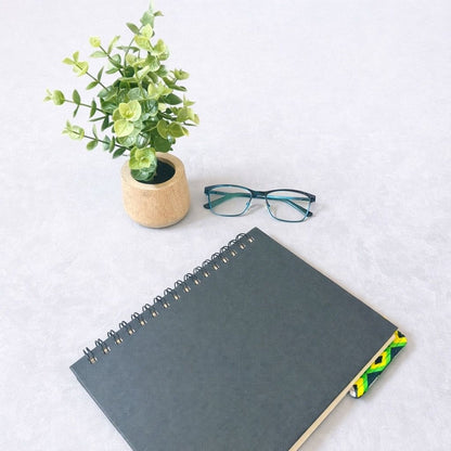 Black notebook with a corner bookmark in black, green, yellow, & white accents on a gray background