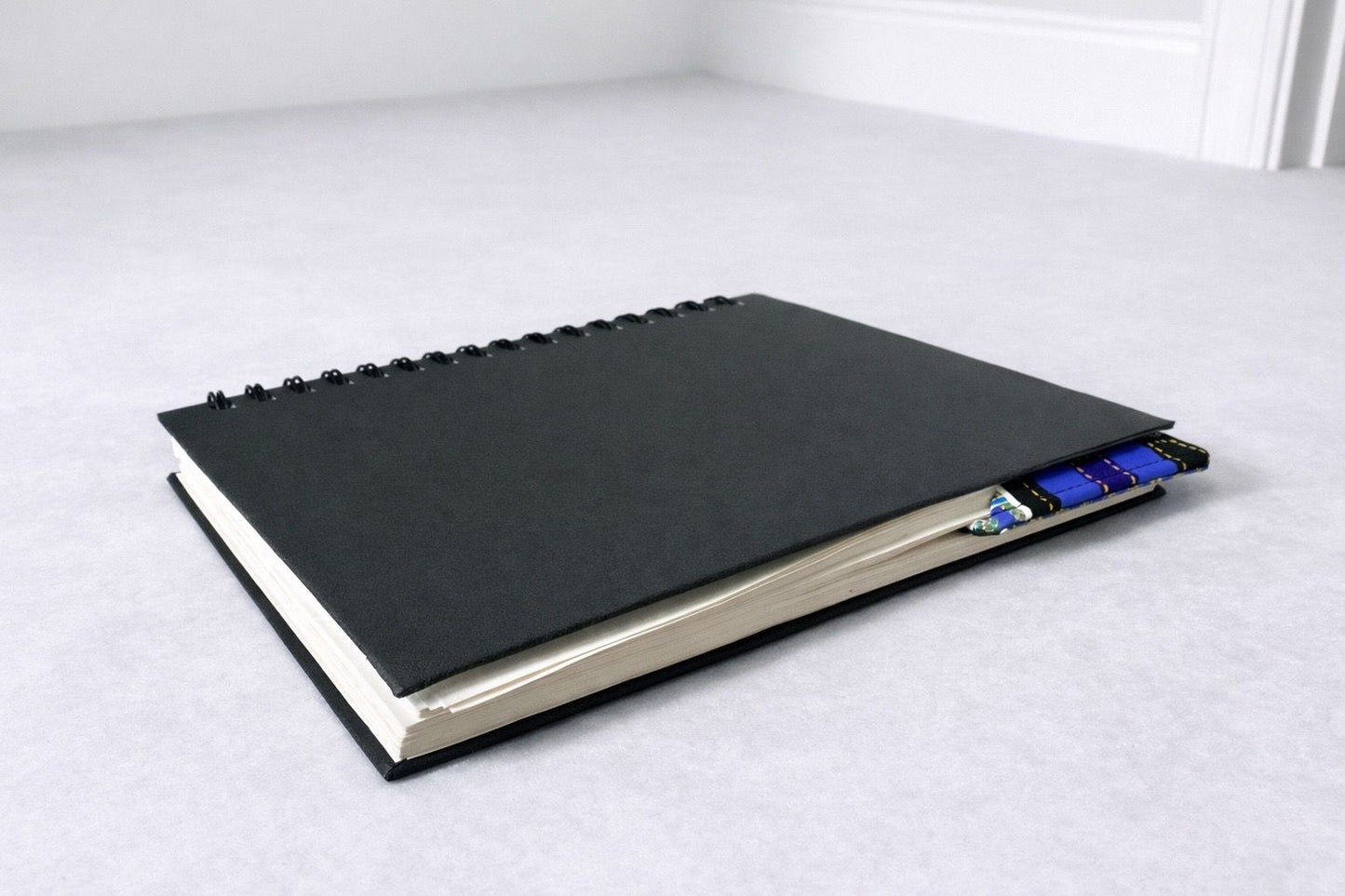 Black notebook with a corner bookmark in blue, dark blue, purple, black, white, & gold accents on a gray background