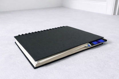 Black notebook with a corner bookmark in blue, dark blue, purple, black, white, & gold accents on a gray background