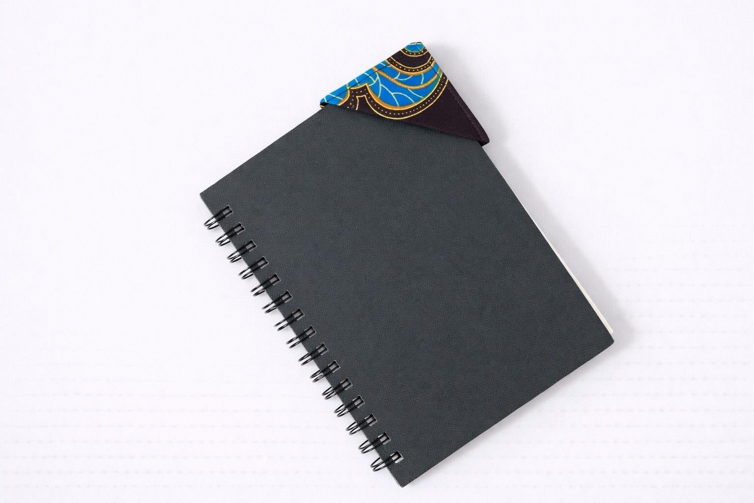 Black notebook with a corner bookmark in dark brown, red, blue, & yellow accents on a gray background