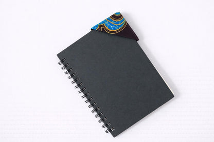 Black notebook with a corner bookmark in dark brown, red, blue, & yellow accents on a gray background