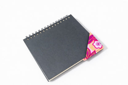 Black notebook with a corner bookmark in pink, dark pink, white, & gold accents on a gray background