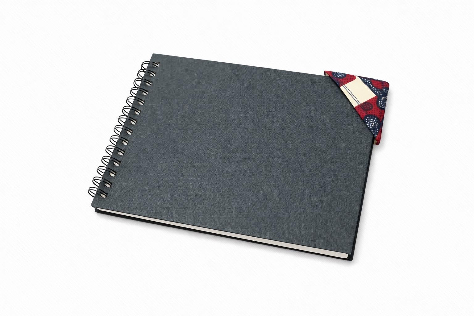 Black spiral-bound notebook with a cream, maroon, & blue corner bookmark on a light gray background