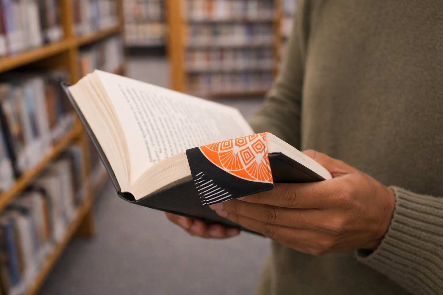 Person holding an open book and a corner bookmark with a library in the background
